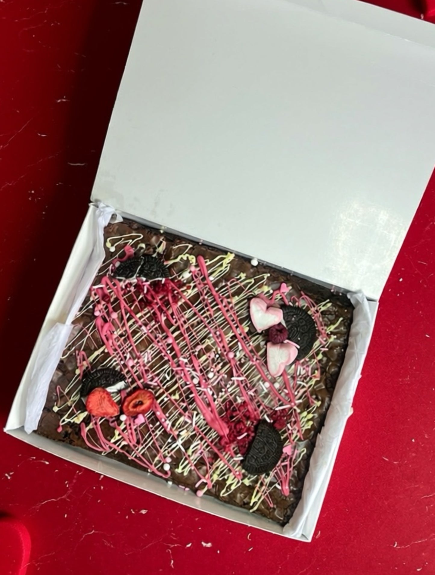Decorative brownie with pink and black drizzle in a white box on a red surface
