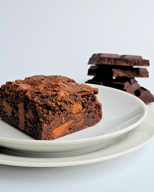 A chocolate brownie on a white plate with chocolate chunks visible, and more chocolate pieces in the background.