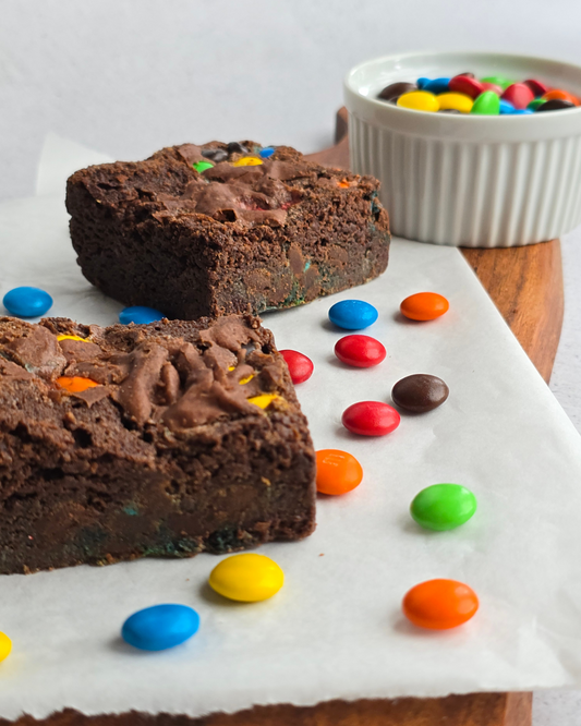 A box of M&M Brownies with one brownie cut open to show the chocolate and M&M toppings, placed on a wooden board with a small white bowl of M&Ms in the background.